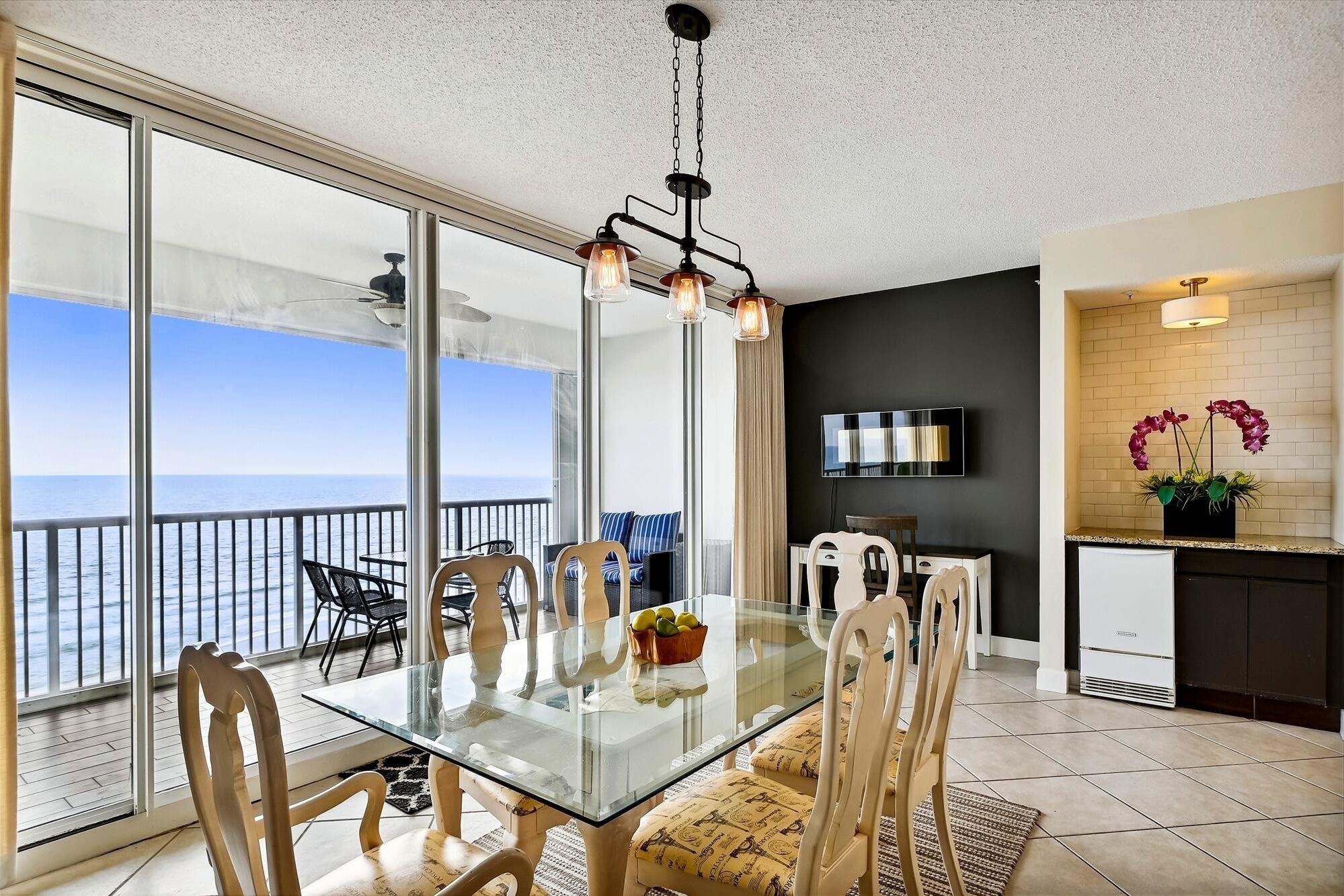 A dining area with a glass table and chairs, a view of the ocean, a kitchen area with a TV, and a potted orchid.