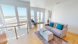 A spacious living room with a large window offering a beach view, a modern sofa with patterned cushions, and a small round table.