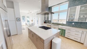 A modern kitchen with a central island, white cabinetry, and a large window offering a view of the beach.