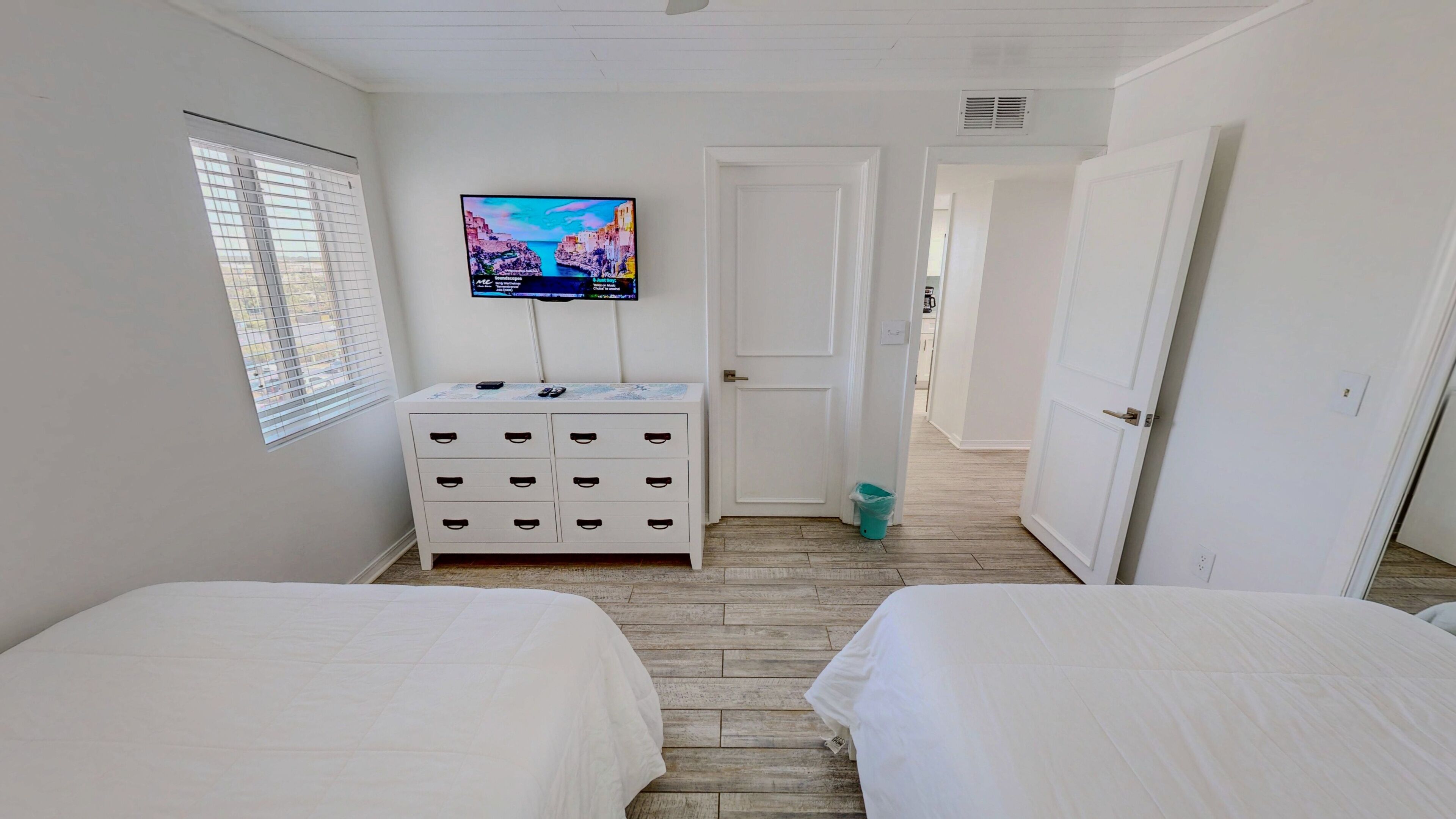 Una camera da letto con due letti, una TV a muro, un comò e una porta che dà su un'altra stanza.