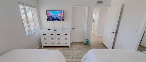 Una camera da letto con due letti, una TV a muro, un comò e una porta che dà su un'altra stanza.