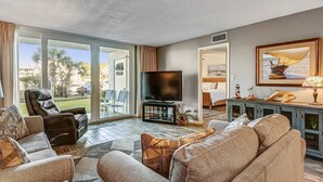 A spacious living room with a leather recliner, a flat-screen TV on a stand, a large sofa, and a dining area visible through a glass door.