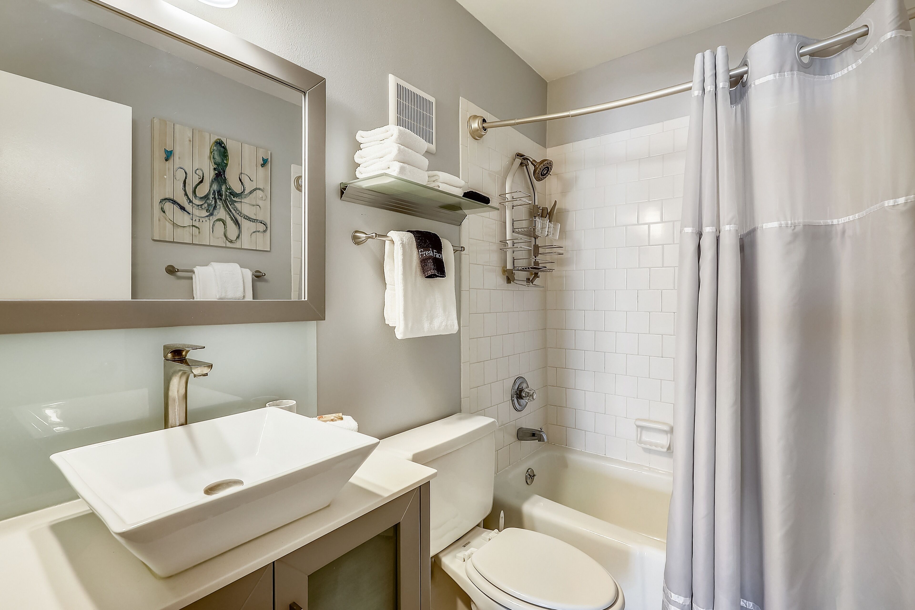 A bathroom with a white sink, toilet, and bathtub. There is a towel rack with towels and a shower curtain. A painting of an octopus is on the wall.