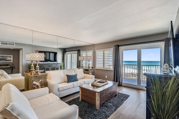 A modern living room with a sofa, coffee table, and a view of the beach through large windows. - Holiday Surf & Racquet 402 2 Bedroom Condo by RedAwning (Destin)