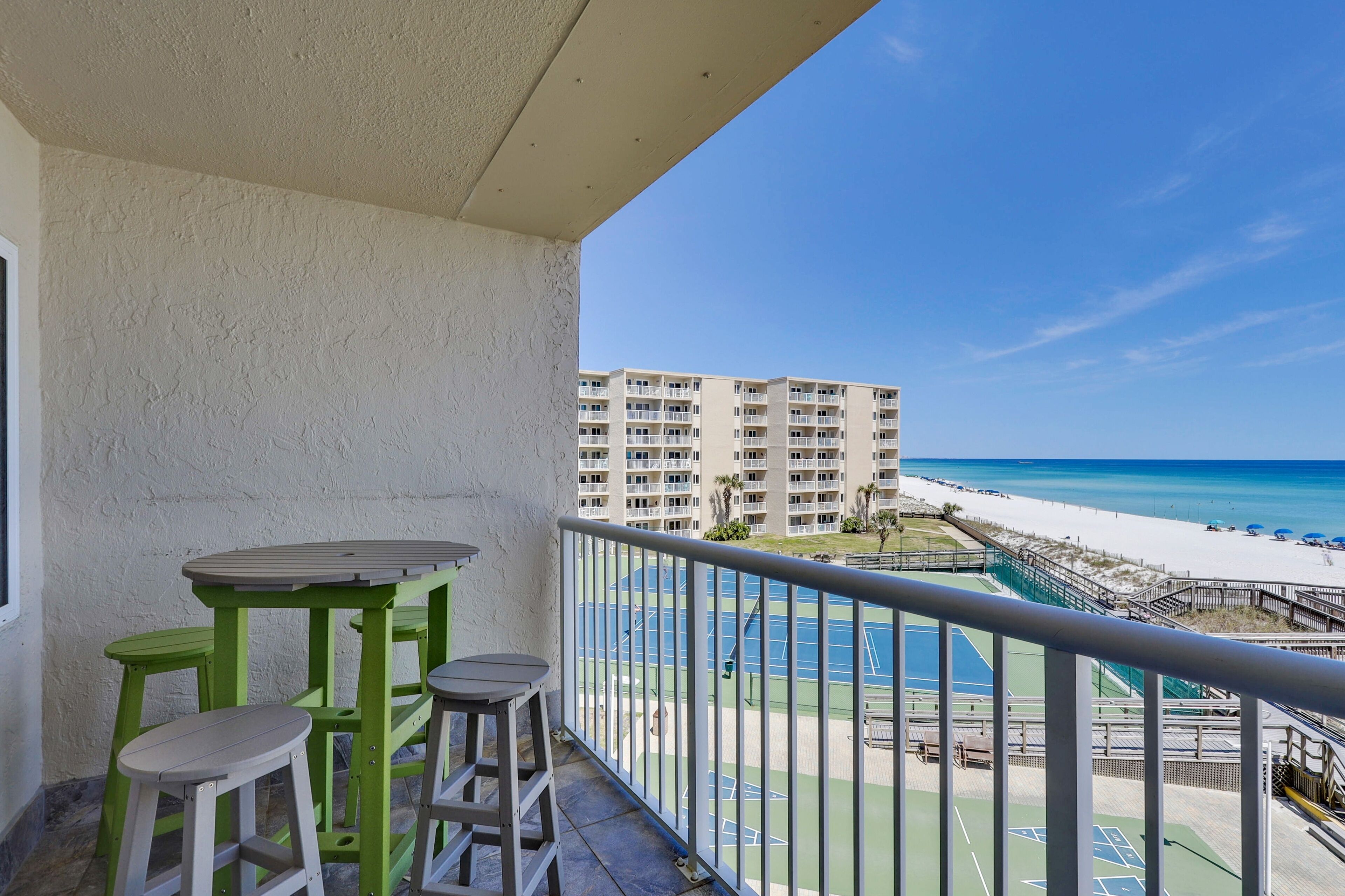 A balcony with a table and two stools, overlooking a beach and a tennis court.