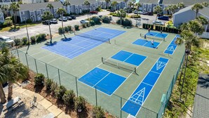 En tennisanläggning med flera banor, omgiven av ett staket, och belägen intill ett bostadsområde med hus och utsikt över stranden.