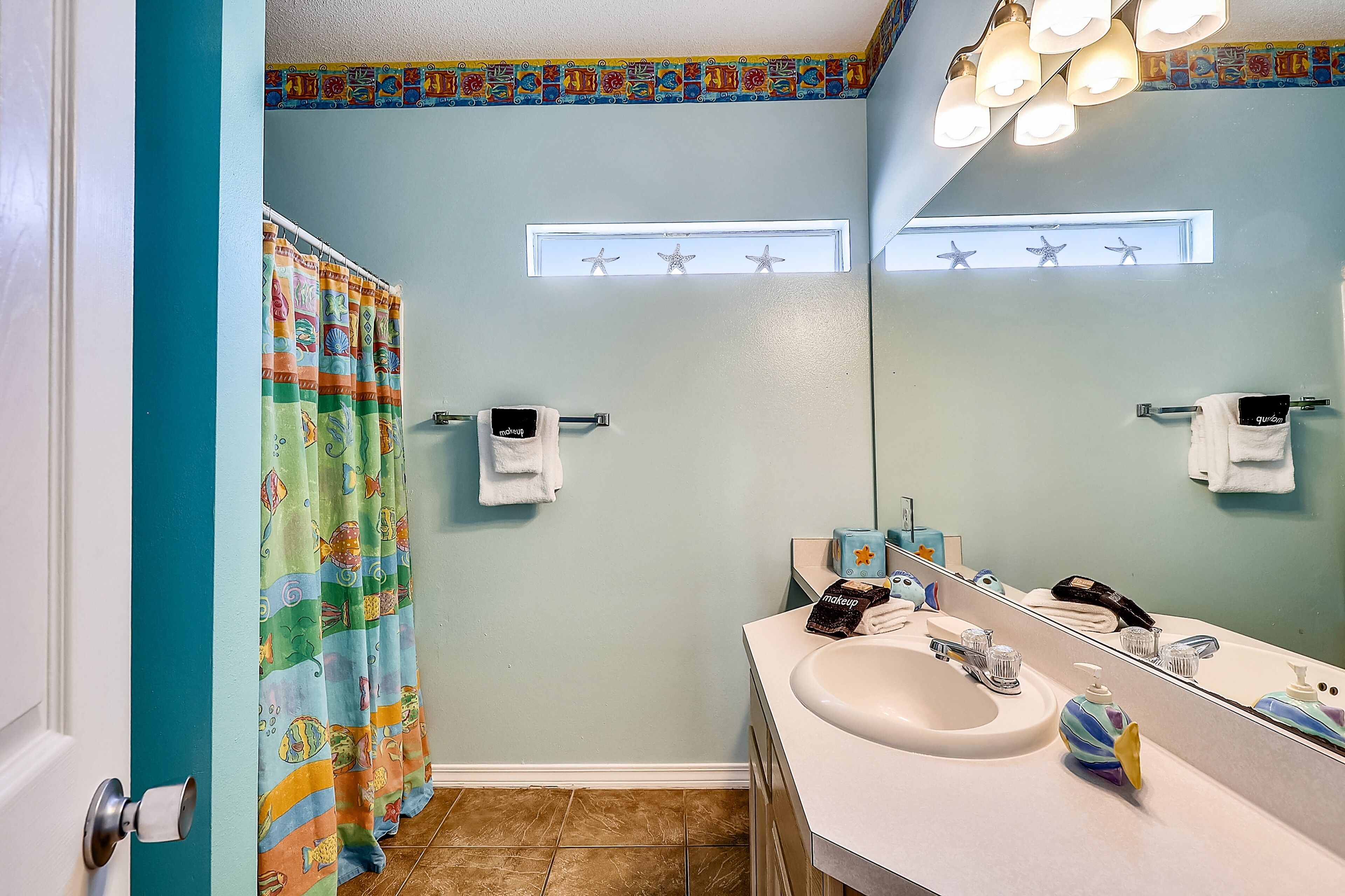 A bathroom with a teal wall, a shower curtain with a colorful pattern, a sink with a mirror, and a ceiling light fixture.