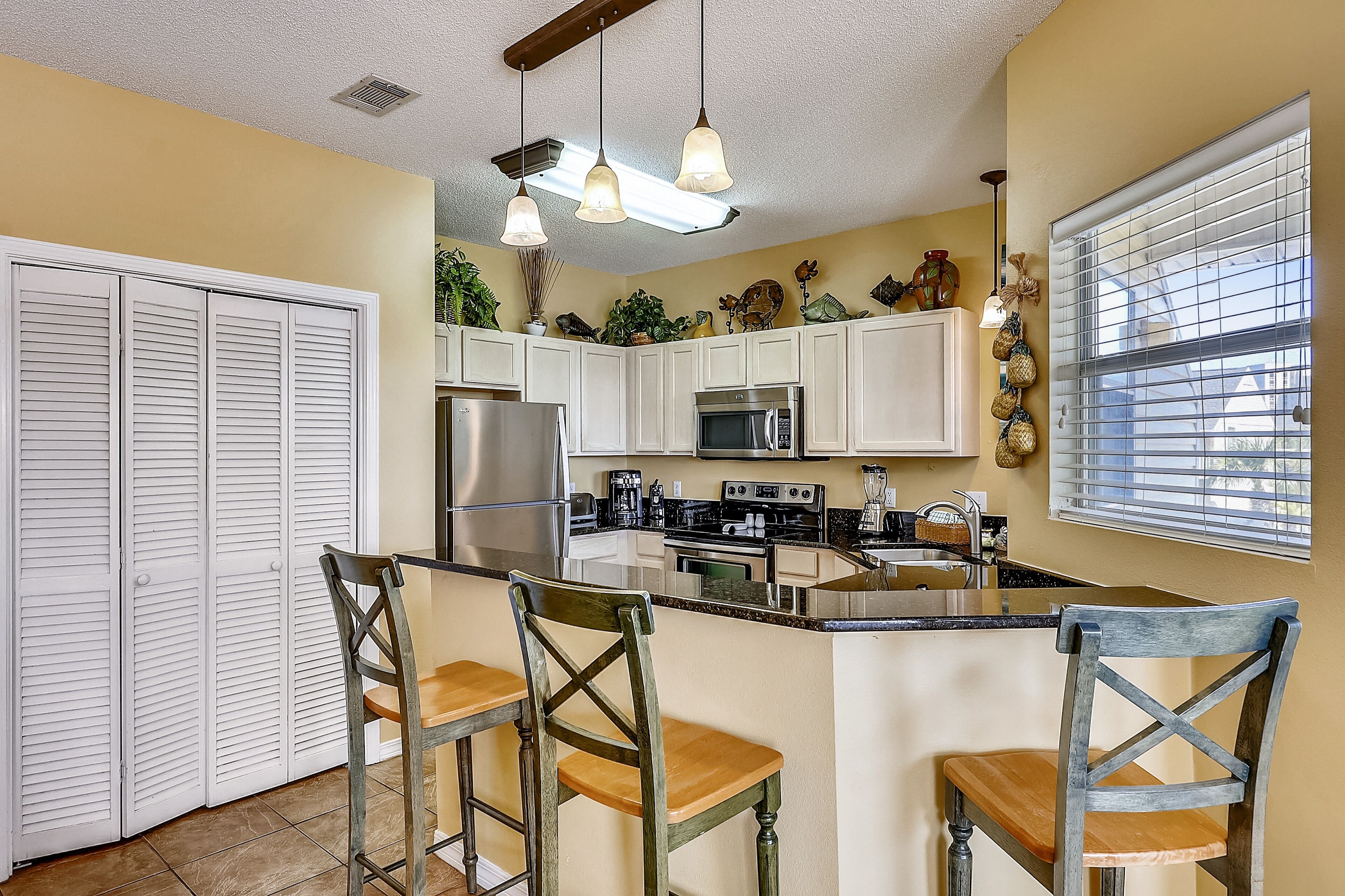 A kitchen with white cabinets, a central island with bar stools, stainless steel appliances, and a window with blinds.