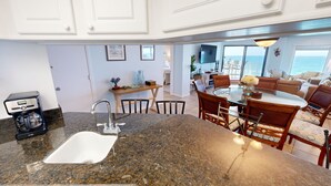 A kitchen with a granite countertop, a coffee maker, and a sink. There is a dining area with a glass table and chairs, and a view of the ocean through large windows.