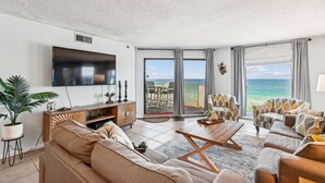 A spacious living room with a large TV, comfortable seating, a coffee table, and a view of the ocean through large sliding doors.