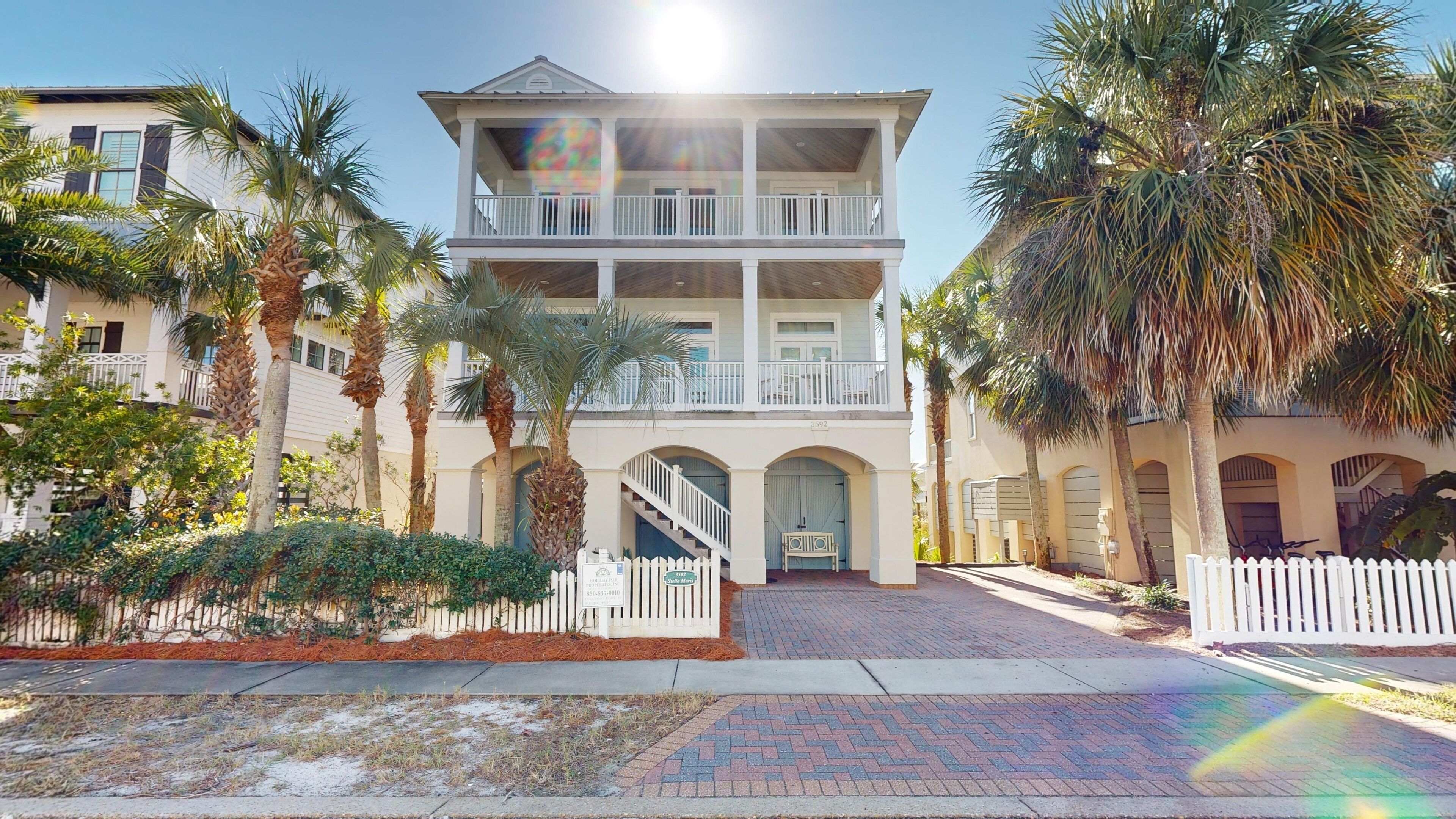 A multi-story building with a balcony, surrounded by palm trees and a white picket fence.