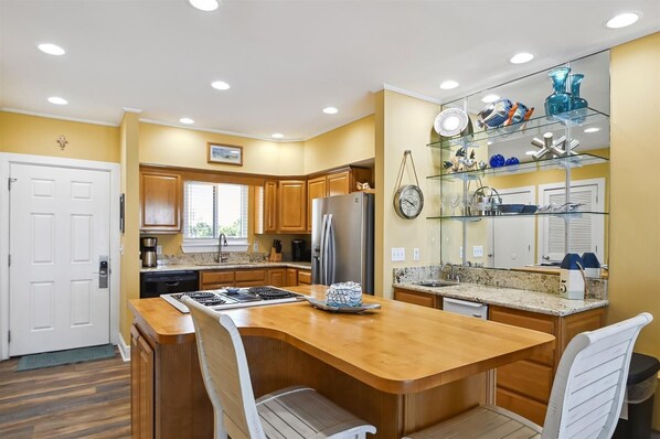 Una cocina con armarios de madera, un refrigerador de acero inoxidable, una encimera de granito y una repisa de vidrio con objetos decorativos.