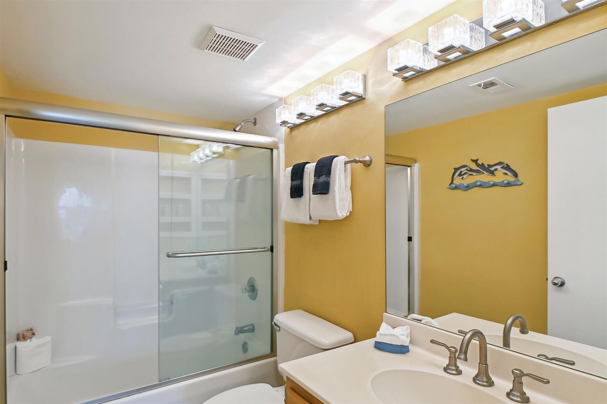 A bathroom with a glass shower enclosure, a toilet, a sink with two faucets, and a mirror with a dolphin decal.