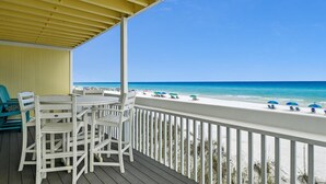 Une terrasse couverte, meublée de mobilier blanc, donnant sur une plage où se trouvent des personnes et des parasols.