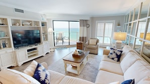 A spacious living room with a large flat-screen TV, comfortable seating, and a view of the ocean through large windows.