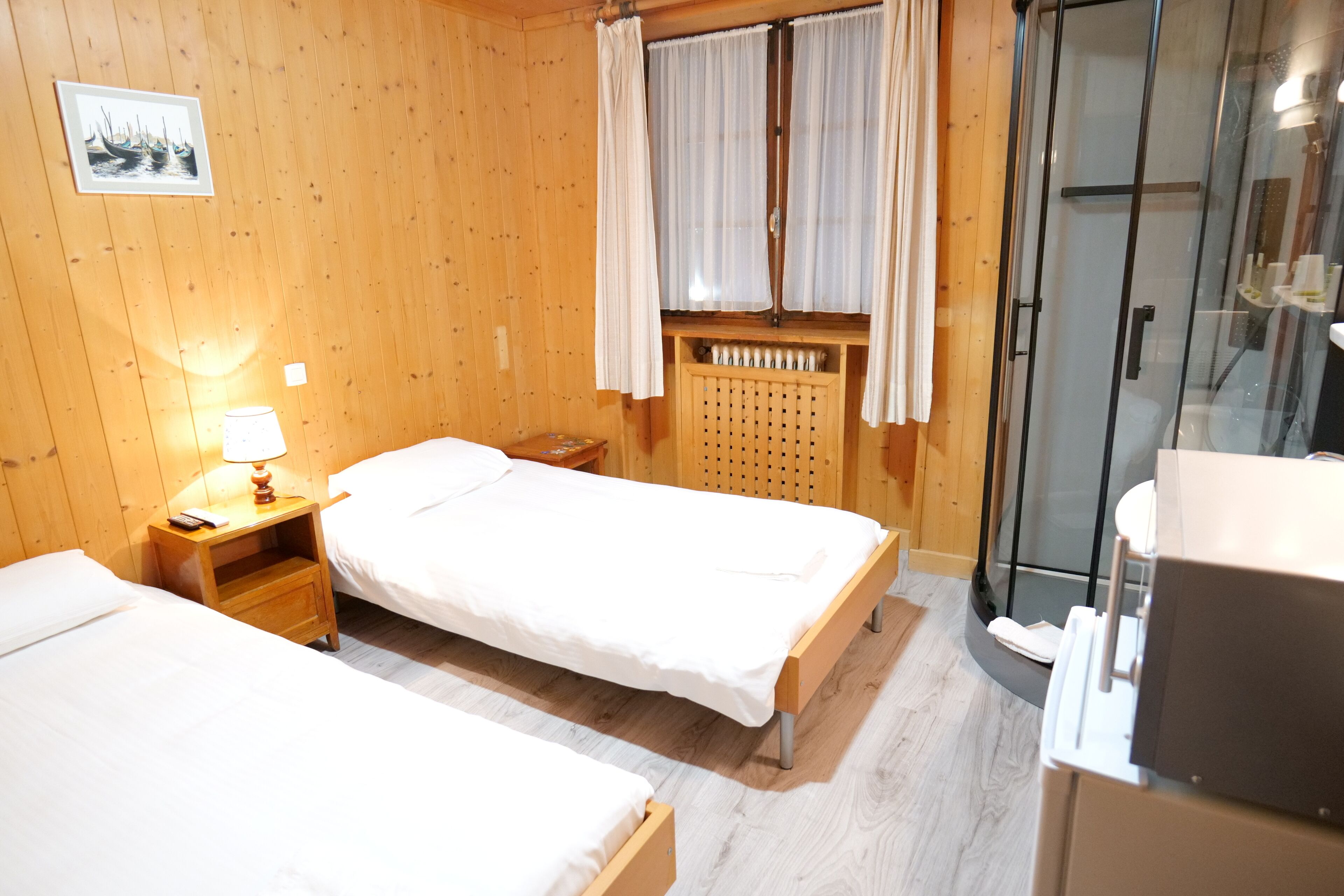 A wooden-paneled room with two beds, a nightstand with a lamp, a window with curtains, and a glass-enclosed shower area.