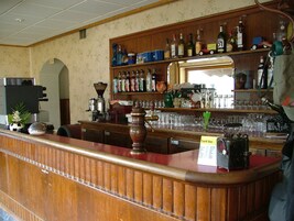 Uma área de bar com balcão de madeira, prateleiras com garrafas, copos e uma placa indicando um bar com preços fixos.