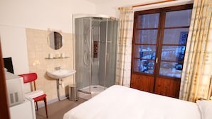 A hotel room with a bed, a shower, a sink, a mirror, and a window with curtains. - Résidence Le Bellevue (Fourneaux)