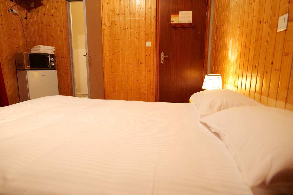 Een hotelkamer met een houten lambrisering, een bed met wit beddengoed, een magnetron, een koelkast en een deur met een bordje.