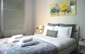 Una cama bien hecha con ropa de cama blanca y una almohada decorativa, una lámpara de noche y un cuadro de flores en la pared.
