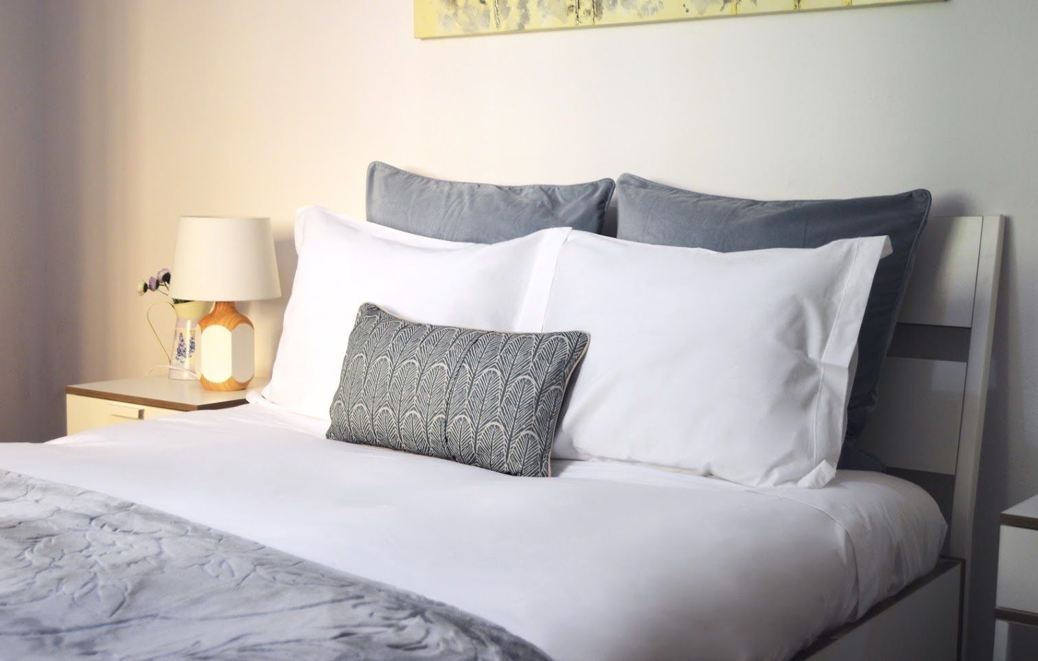 A neatly made bed with white bedding and a patterned pillow, a bedside lamp, and a small vase with flowers on a nightstand.