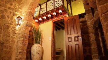 Um corredor de paredes de pedra com uma porta de madeira, um vaso grande com plantas e iluminação decorativa.