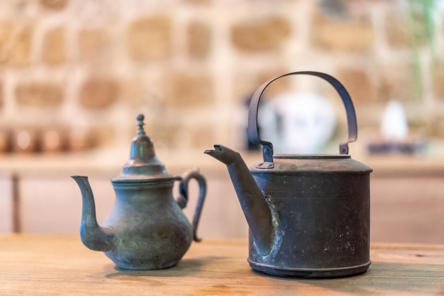 Two metal teapots on a wooden surface with a stone wall in the background.
