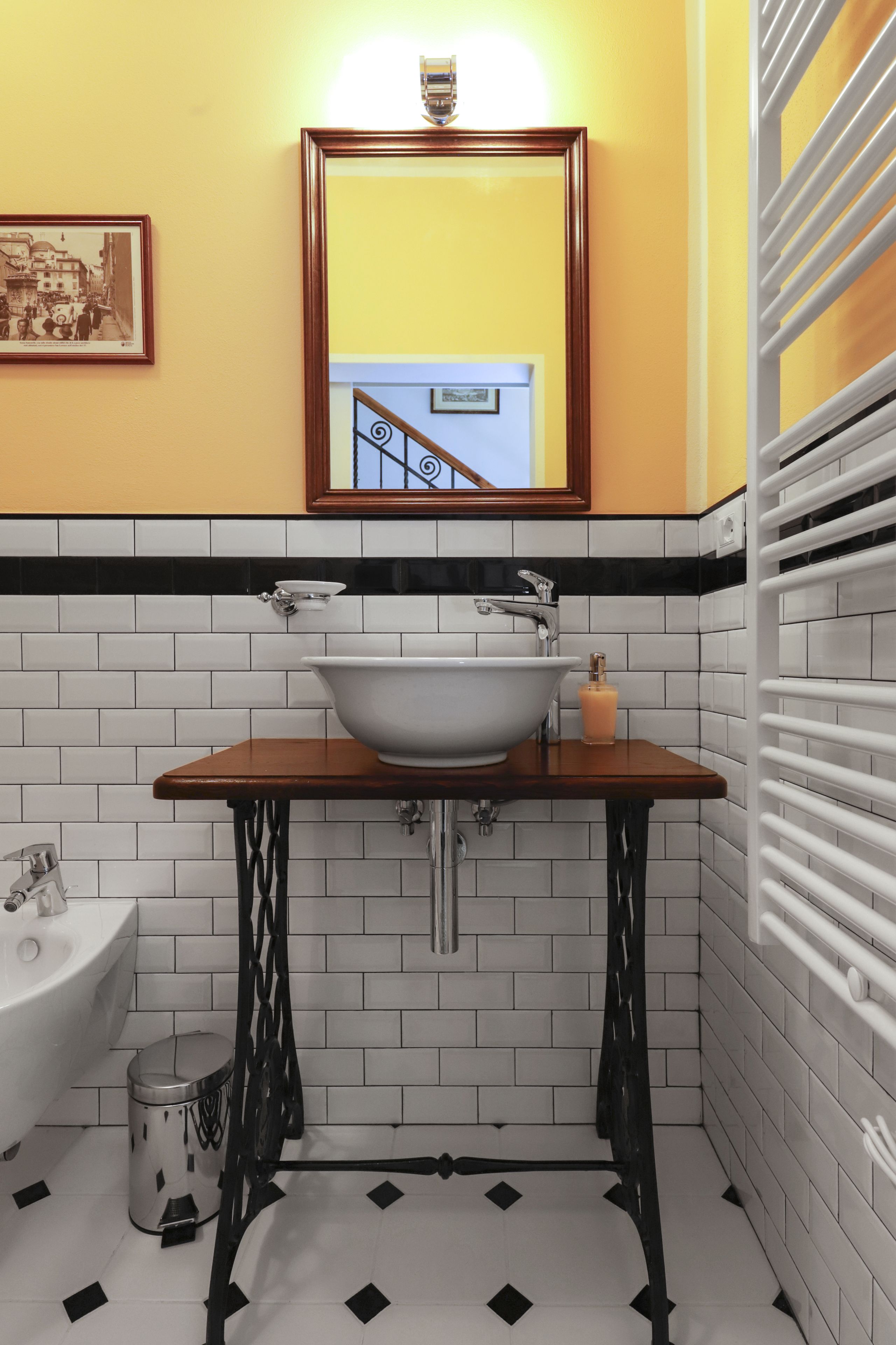 Een badkamer met een wit wastafel, zwart-wit tegels, een houten wastafelmeubel en een spiegel met een gele muur erachter.