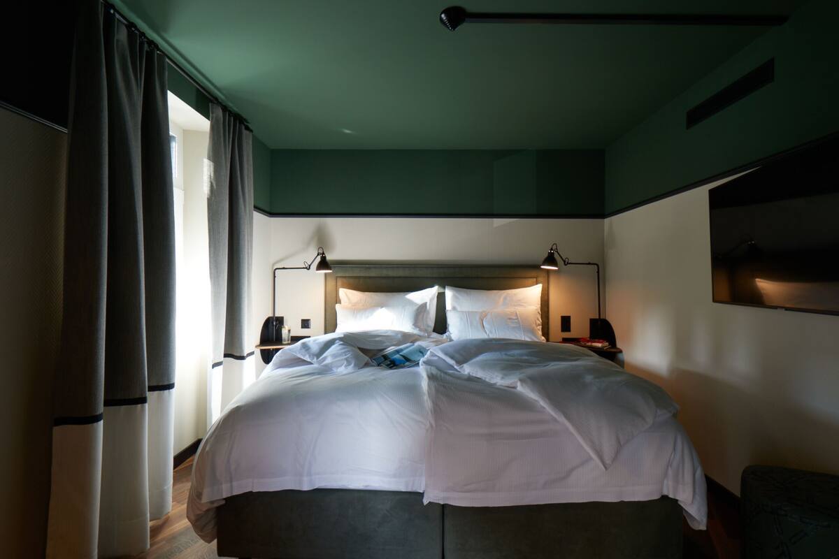 A neatly made bed with white linens, a bedside table with a lamp, and a window with curtains.