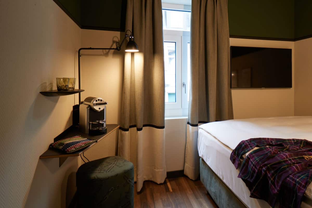 A bedroom with a bed, a desk with a lamp, a coffee maker, and a window with curtains.