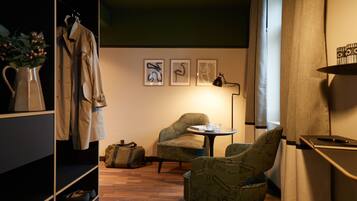 Ein Hotelzimmer mit Holzboden, einer Sitzecke mit zwei Stühlen, einem kleinen Tisch mit Tasse und Buch, einem Kleiderhaken mit Jacke und einer Wand mit drei gerahmten Bildern.