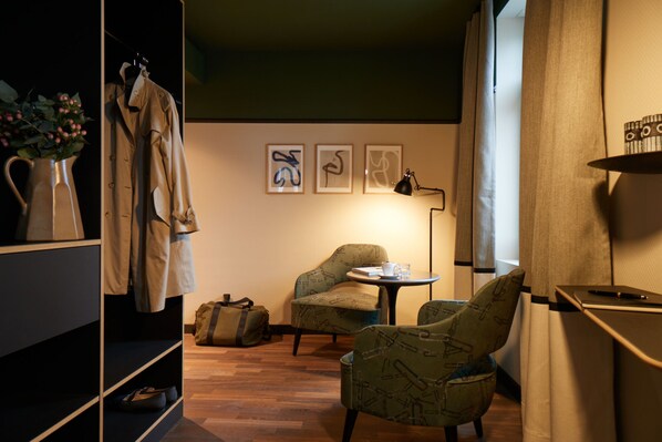 A hotel room with a wooden floor, a seating area with two chairs, a small table with a cup and a book, a coat rack with a jacket, and a wall with three framed pictures.