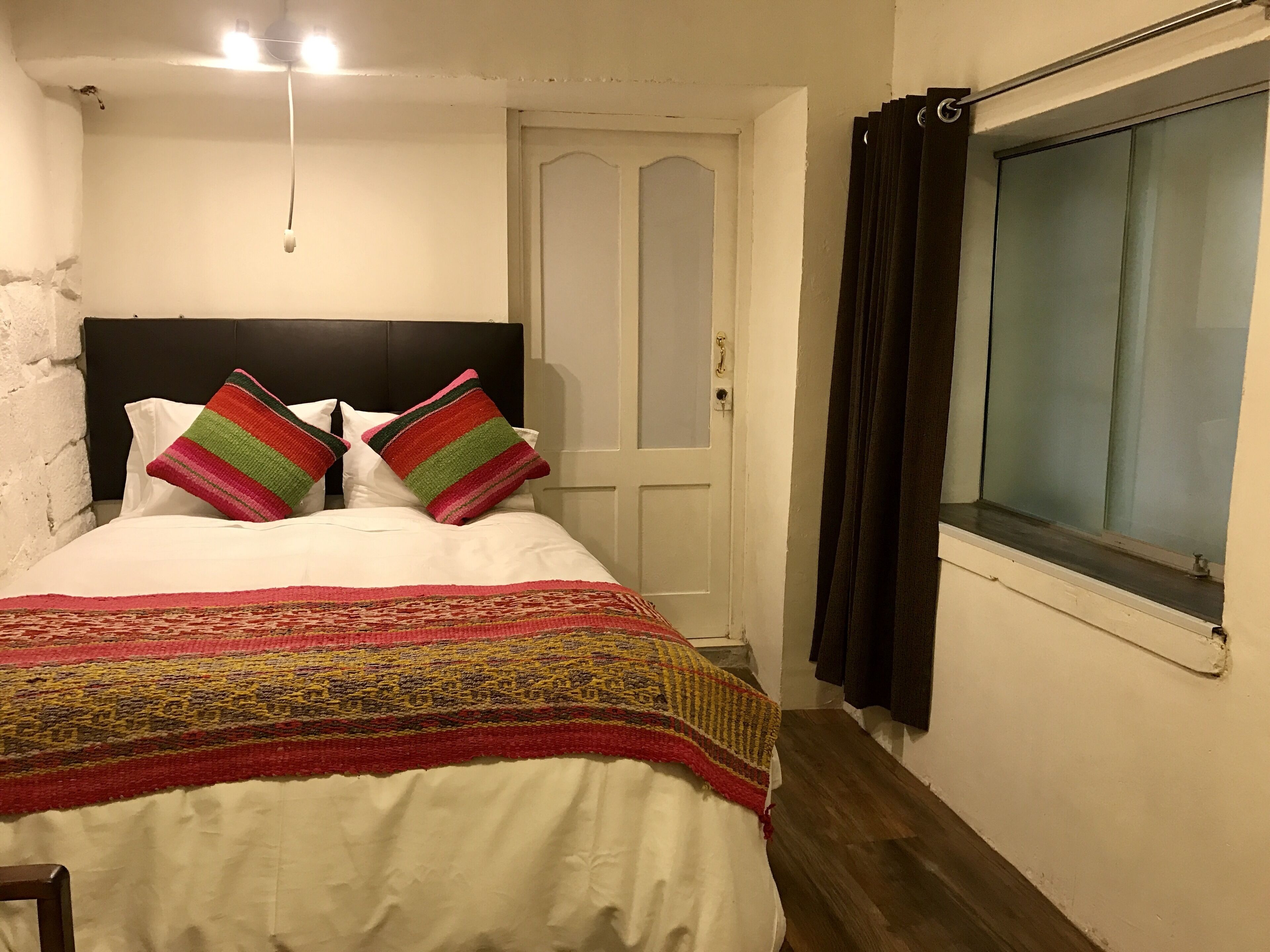 A bedroom with a bed, a headboard, a door, a window with curtains, and a wall with a light fixture.
