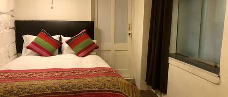 A bedroom with a bed, a headboard, a door, a window with curtains, and a wall with a light fixture.