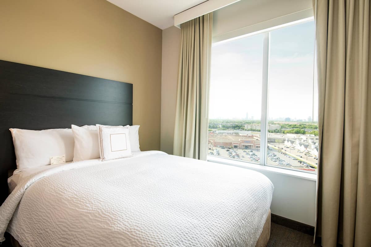 A hotel room with a large bed, a dark headboard, and a view of a cityscape through a large window.