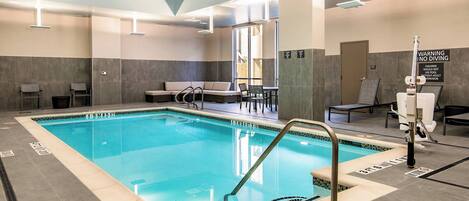 An indoor swimming pool with clear lane markings, a changing room area, and a seating area with a view of the outdoors.