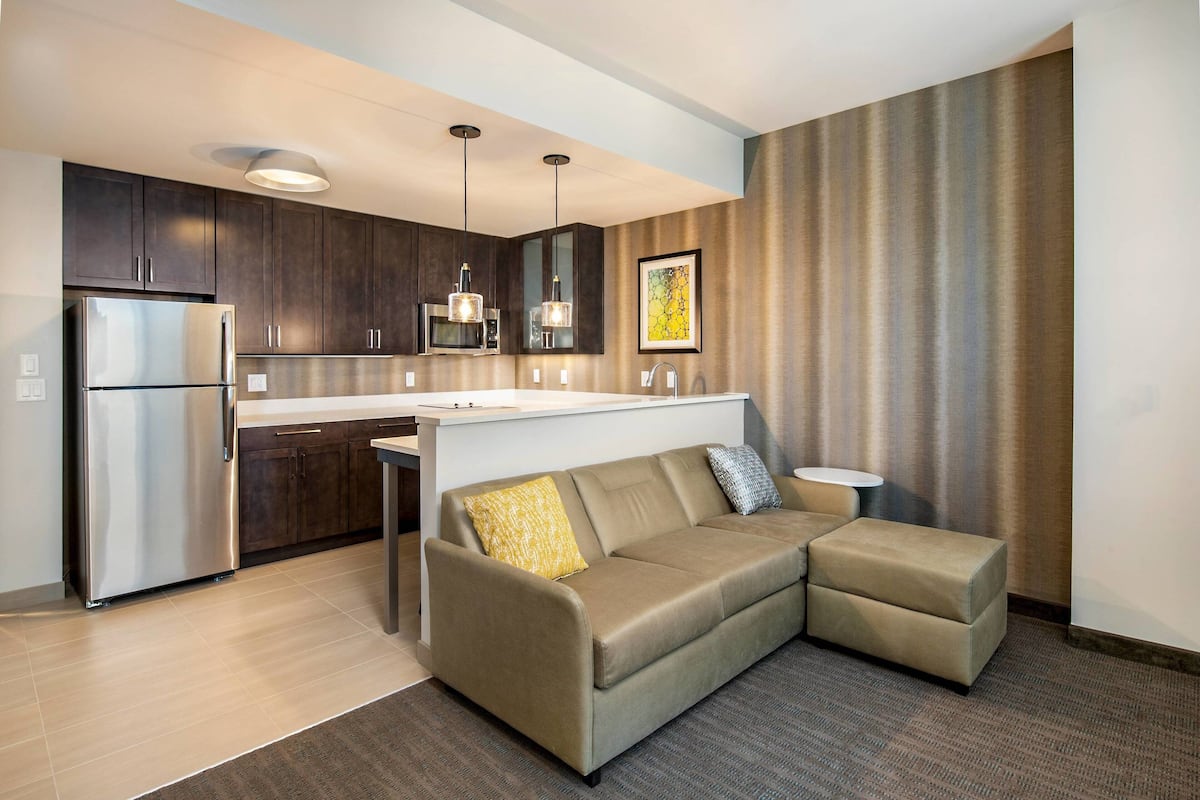 A modern kitchen with dark cabinets, a stainless steel refrigerator, pendant lights, and a small island with a sink.