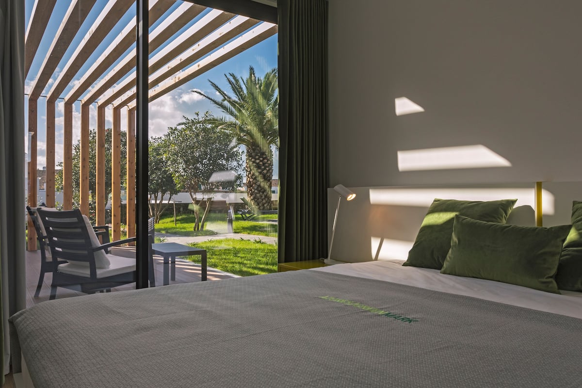 A modern hotel room with a large bed, a view of the outdoors, and a balcony with seating.