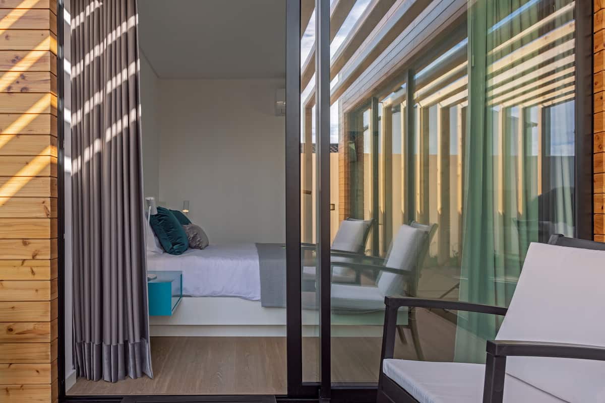A modern room with a bed, a small table, and a chair. There is a sliding glass door leading to an outdoor area with a table and chairs.