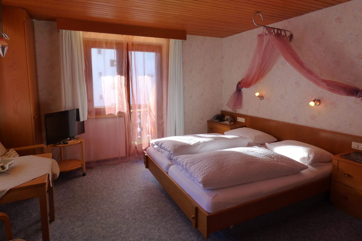 A bedroom with a wooden bed, a bedside table, a TV on a small stand, and a window with sheer curtains.