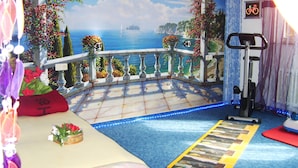 Une pièce ornée d’une fresque représentant un balcon donnant sur la mer, avec un vélo d’appartement et une petite plante posée sur le sol.