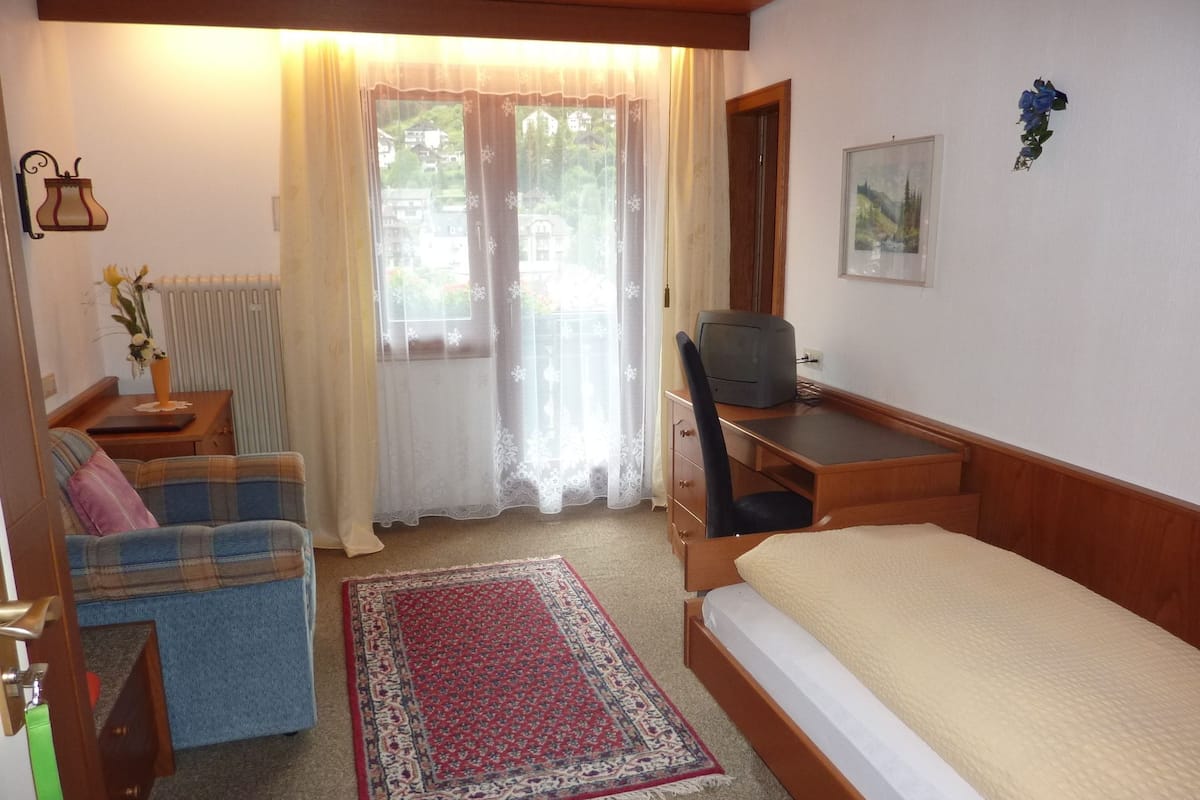 A small, cozy room with a bed, a desk, a chair, a sofa, and a window with curtains.