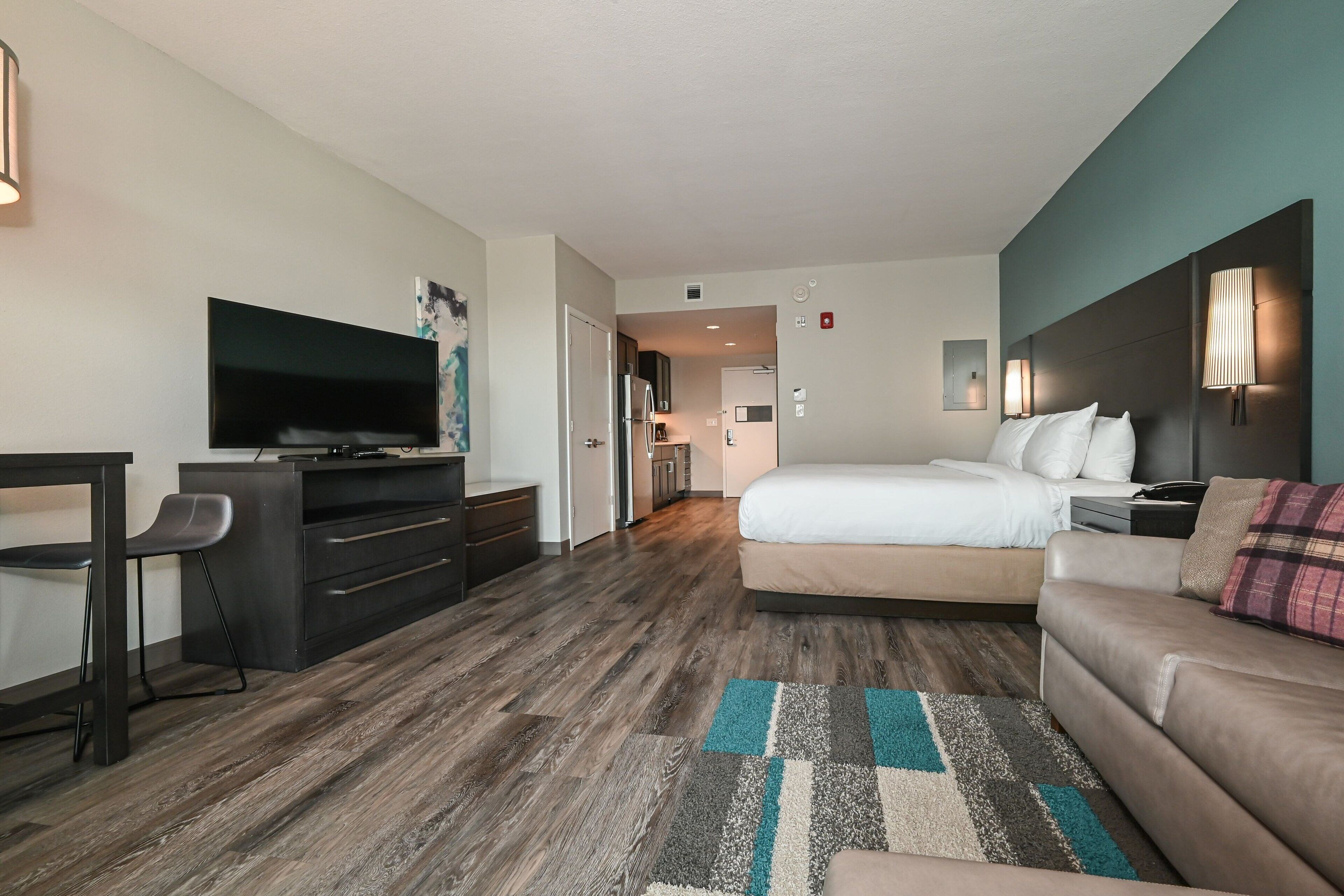 Quarto de hotel moderno com televisão de tela plana, escrivaninha, cama confortável, sofá e cozinha ao fundo.