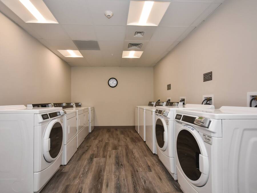 A laundry room with four white front-loading washers and dryers, wooden flooring, and a clock on the wall.