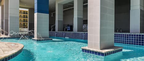 An indoor swimming pool with clear blue water, surrounded by white and blue tiled walls, and supported by large pillars.
