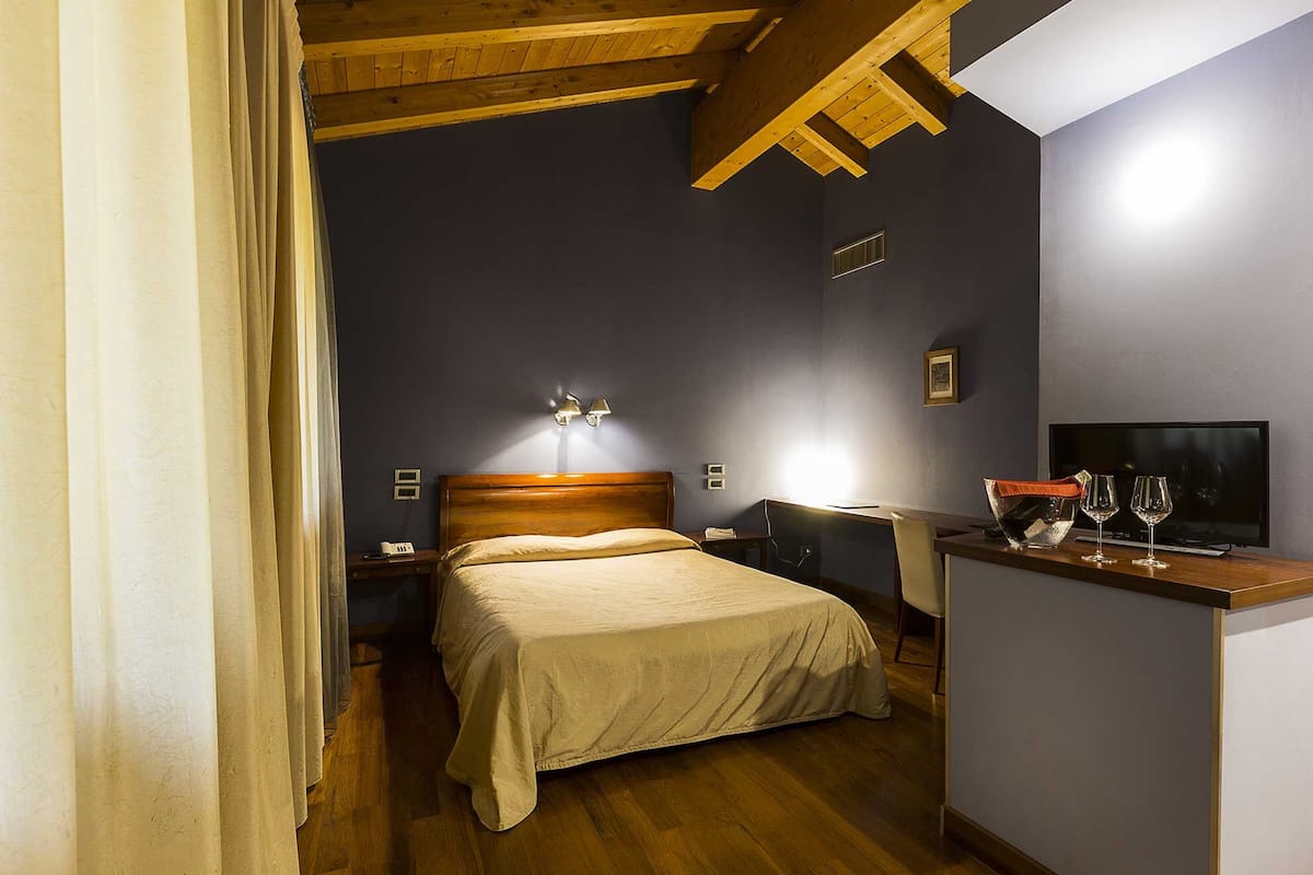 A hotel room with a wooden ceiling, a bed with a headboard, bedside lamps, a desk with a chair, and a flat-screen TV.