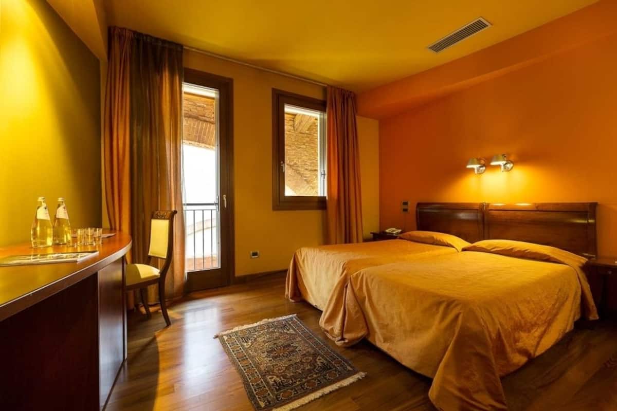 A hotel room with a double bed, a desk with a chair, a window with curtains, and a nightstand with a lamp.