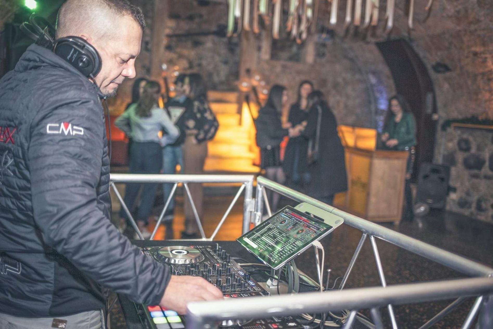 A DJ booth with a control panel and a laptop, set against a stone wall with a crowd in the background.