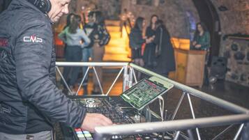 A DJ booth with a control panel and a laptop, set against a stone wall with a crowd in the background.
