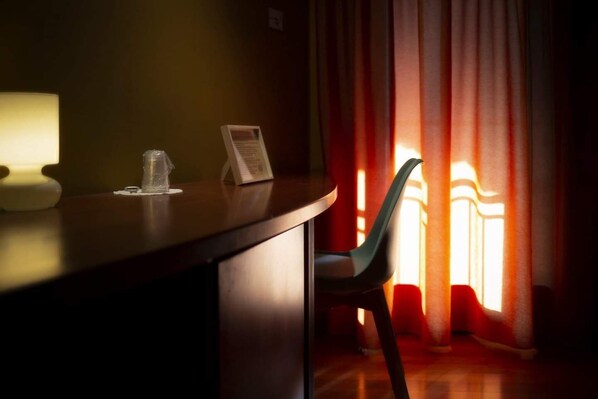 Una habitación tenuemente iluminada con un escritorio de madera, una silla y un ventanal con cortinas rojas.